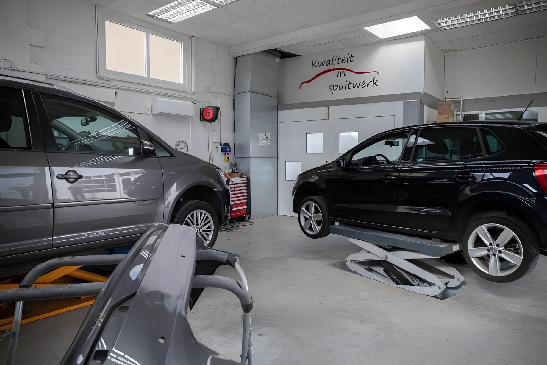De werkplaats met twee auto's op heftafels en de spuitcabine op de achtergrond