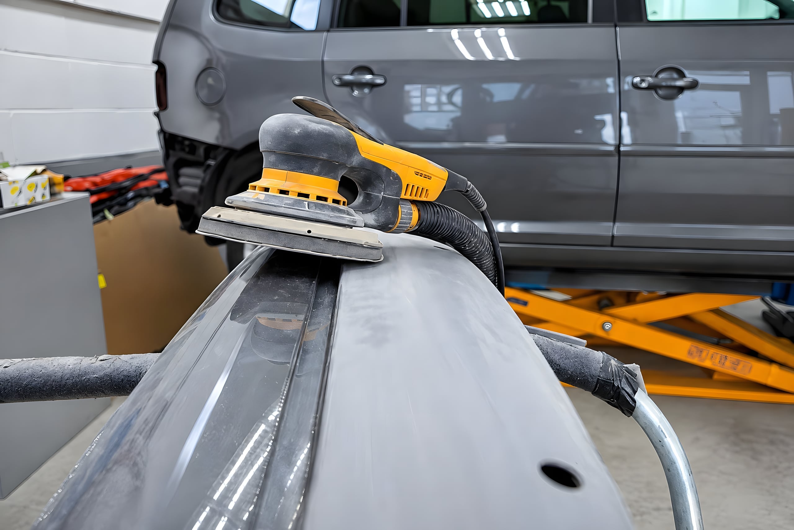 Excentrische schuurmachine op een gedemonteerde bumper in de werkplaats, met een auto op de heftafel erachter